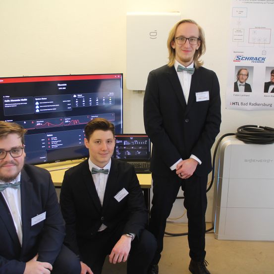 Drei junge Männer in Anzügen und Fliegen stehen vor einem Computerbildschirm. Der Mann auf der rechten Seite trägt eine Brille.