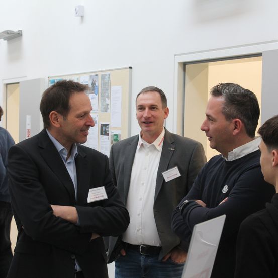 Drei Männer lächeln und unterhalten sich in einem weiß getünchten Raum. Sie tragen formelle Kleidung, und einer von ihnen trägt einen Namensschild.