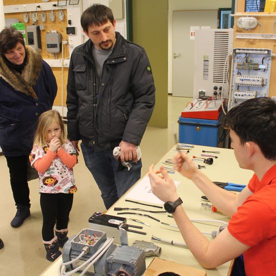 Eine Gruppe von Menschen ist in einem Raum mit Werkzeugen und Ausrüstung. Ein Mann zeigt einer kleinen Mädchen, wie man ein Werkzeug benutzt.