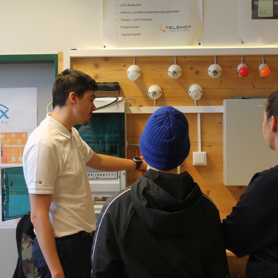 Drei Personen stehen vor einer Wand mit verschiedenen Geräten. Der Mann im weißen Hemd zeigt auf etwas. Die Frau rechts trägt eine schwarze Jacke.