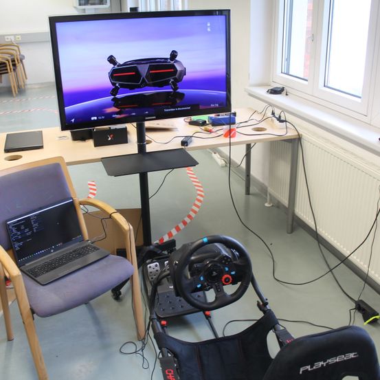 Ein Gaming-Setup mit Monitor, Lenkrad und Laptop auf einem Stuhl in einem Raum mit Fenstern und Heizkörpern.