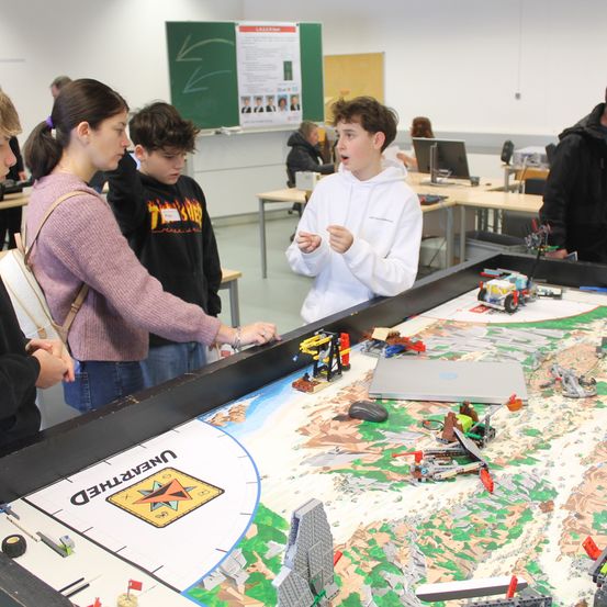 Eine Gruppe von Schülern steht um einen großen Tisch herum, auf dem eine Lego-Kreation ausgestellt ist. Ein Schüler spricht aufgeregt, während andere die detaillierte Einrichtung betrachten.