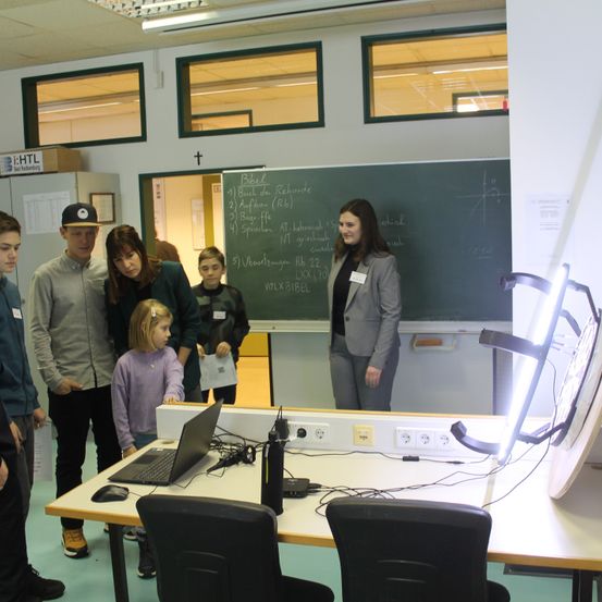 Eine Gruppe von Menschen in einem Klassenzimmer mit einer Tafel und einem Laptop auf einem Tisch. Eine Frau steht vor der Tafel, während andere den Laptop betrachten.