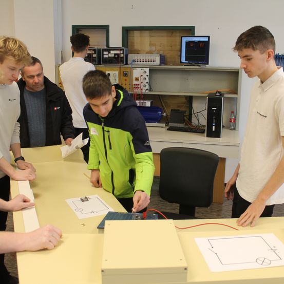 Eine Gruppe junger Menschen arbeitet in einem Klassenzimmer an einer Leiterplatte. Auf dem Tisch liegen Papiere mit Zeichnungen.