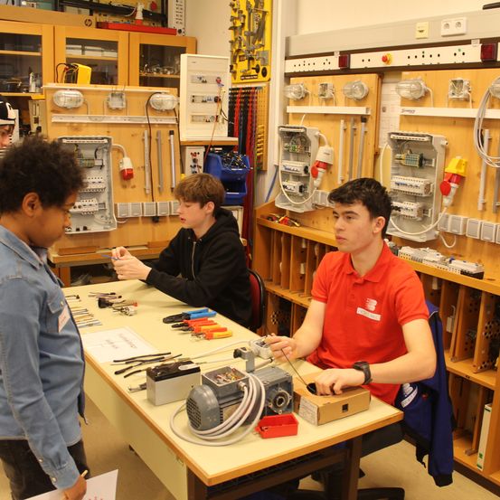 Drei Schüler sind in einem Klassenzimmer und arbeiten an einem elektrischen Projekt. Der Mann im roten Hemd sitzt an einem Tisch mit einem Motor, während die anderen in ein Gespräch vertieft sind.