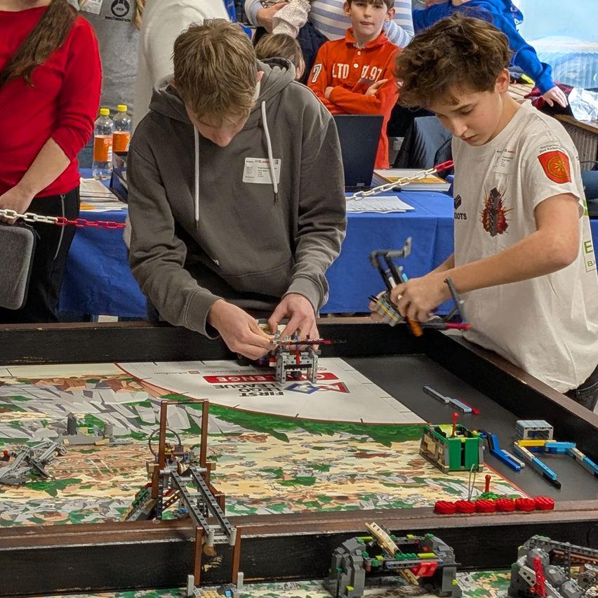 Zwei junge Jungen bauen mit Legos auf einem Tisch bei einem Wettbewerb. In der Nähe beobachten Erwachsene und Kinder.