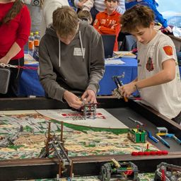 Zwei junge Jungen bauen mit Legos auf einem Tisch bei einem Wettbewerb. In der Nähe beobachten Erwachsene und Kinder.