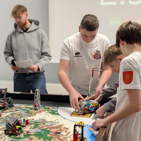Drei Jungen bauen mit Lego, einer in einer Kapuzenjacke und zwei in weißen T-Shirts. Sie sind in einem Raum mit einer Leinwand dahinter.