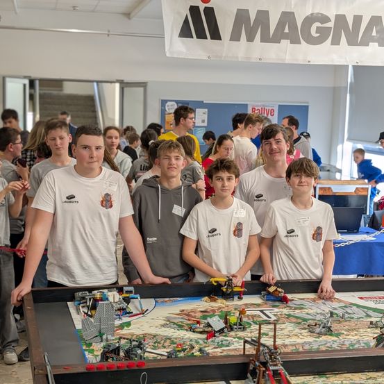 Eine Gruppe junger Menschen in übereinstimmenden weißen T-Shirts steht an einem großen Lego-Tisch. Dahinter ist eine Menschenmenge, ein Banner mit dem Wort Magna und eine Treppe.