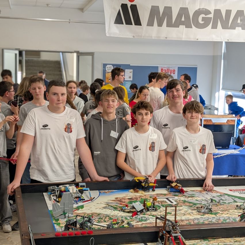 Eine Gruppe junger Menschen in übereinstimmenden weißen T-Shirts steht an einem großen Lego-Tisch. Dahinter ist eine Menschenmenge, ein Banner mit dem Wort Magna und eine Treppe.