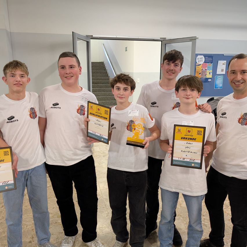 Eine Gruppe junger Jungen und Männer, die weiße T-Shirts tragen, stehen zusammen in einem Raum und lächeln für ein Foto. Sie halten Urkunden in der Hand und einer von ihnen trägt eine Medaille. Hinter ihnen befindet sich eine Wand mit einer Pinnwand.