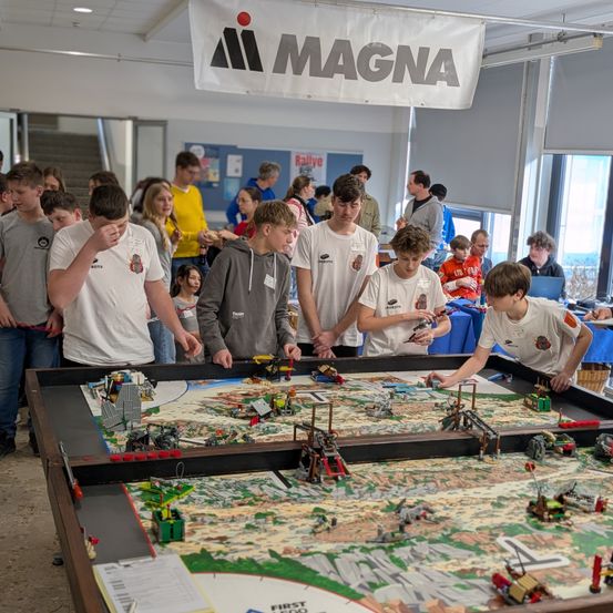Eine Gruppe junger Leute in weißen T-Shirts versammelt sich um einen großen Tisch mit einer LEGO-Karte und -Modellen. Der Tisch befindet sich in einem Raum mit einem Banner, das 'MAGNA' und Fenster zeigt.