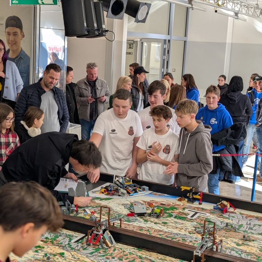 Eine Gruppe von Menschen, darunter Jungen und Mädchen, hat sich um einen Tisch mit einer Lego-Ausstellung versammelt. Ein Mann beugt sich vor und schreibt auf ein Blatt Papier. Der Tisch hat verschiedene Lego-Kreationen. Einige Leute beobachten die Ausstellung.