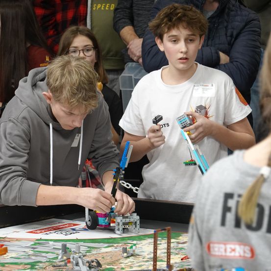 Zwei Jungen bauen Lego-Roboter auf einem Tisch vor einem Publikum zusammen. Ein Junge trägt eine graue Kapuzenjacke und der andere ein weißes T-Shirt mit Lego-Marke.