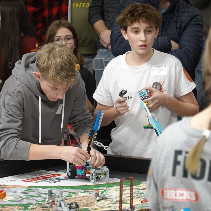 Zwei Jungen bauen Lego-Roboter auf einem Tisch vor einem Publikum zusammen. Ein Junge trägt eine graue Kapuzenjacke und der andere ein weißes T-Shirt mit Lego-Marke.