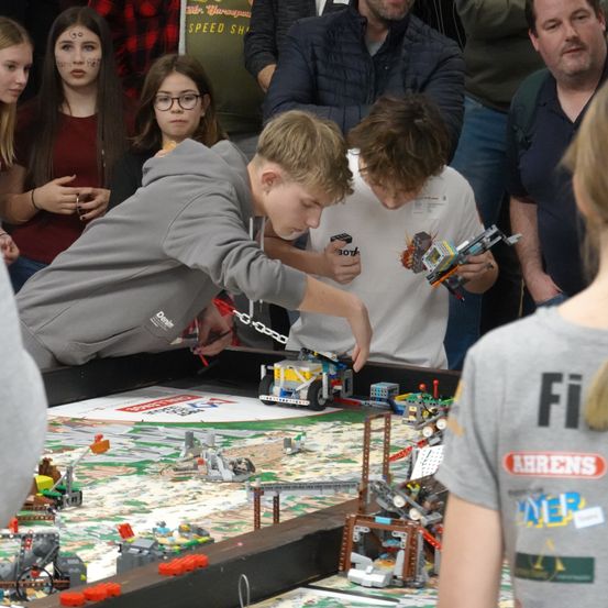 Eine Gruppe junger Jungen spielt mit Legos auf einem großen Tisch mit grünem Hintergrund. Sie werden von Erwachsenen begleitet, die zuschauen.