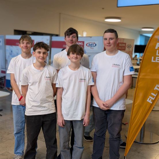 Fünf Jungen stehen in einer Reihe und lächeln, einer trägt ein rotes Armband. Sie tragen alle weiße T-Shirts. Hinter ihnen befindet sich ein orangefarbener Banner mit weißem Text.