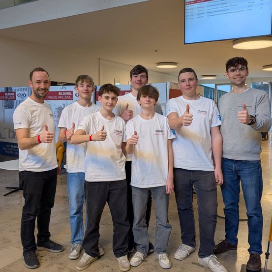 Eine Gruppe junger Jungen und Männer in weißen T-Shirts, die zusammenstehen und in einem Raum Daumen hoch geben. Die beiden Erwachsenen lächeln.