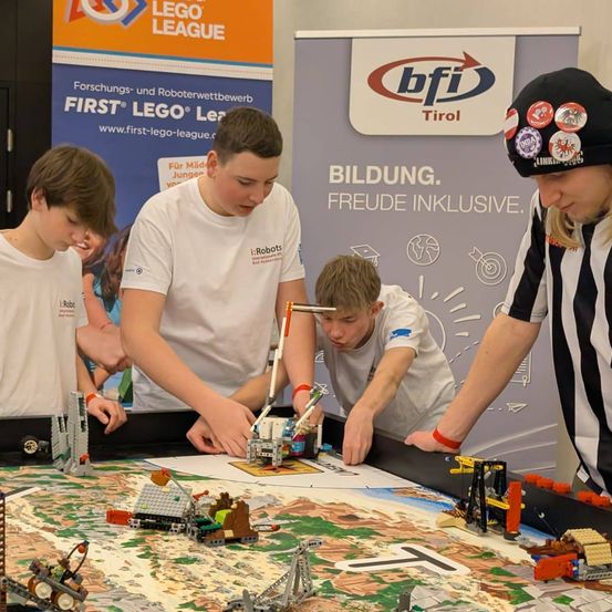 Mehrere Kinder arbeiten an einem Tisch mit verschiedenen Lego-Robotern. Ein Kind konzentriert sich auf die Roboter, während ein anderes zusieht. Eine Frau in einer Schiedsrichteruniform beobachtet. Hinter ihnen ist eine Wand mit einem FIRST Lego League-Banner.