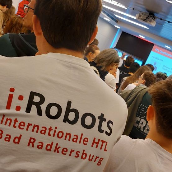 Eine Gruppe von Menschen sitzt in einem Raum, einige tragen Brillen. Ein Mann vorne trägt ein weißes T-Shirt mit schwarzem Text, der 'i:Robots' steht. Eine Projektionsleinwand zeigt eine Nachricht an der Wand.