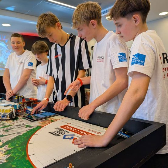 Eine Gruppe junger Jungen in weißen T-Shirts spielt mit Lego-Robotern auf einem Tisch. Ein Junge trägt ein schwarz-weiß gestreiftes Shirt.