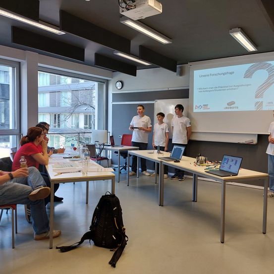 Eine Gruppe von Studenten sitzt um einen Tisch herum mit Laptops und anderem Equipment. Ein Beamer zeigt Text dahinter. Zwei Männer stehen vor dem Tisch.