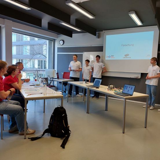 Eine Gruppe von Menschen befindet sich in einem Besprechungsraum mit Tischen und Laptops. Eine Präsentation läuft auf einer Projektionsleinwand. Es sind Schüler und ein Lehrer anwesend.