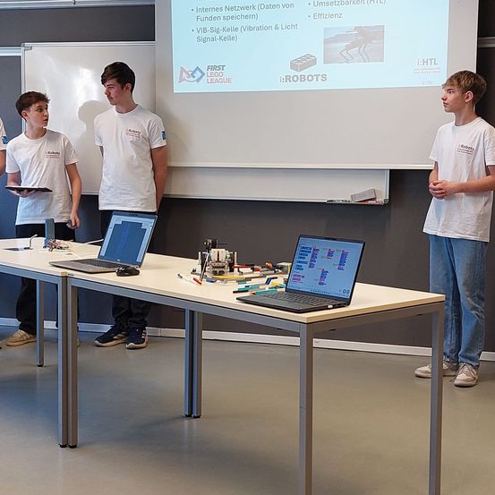 Vier junge Männer stehen um einen Tisch mit Laptops herum und blicken auf eine Projektionsleinwand mit einer Präsentation mit dem Titel 'FIRST LEGO LEAGUE.' Sie tragen weiße Hemden mit Logos und Turnschuhen.