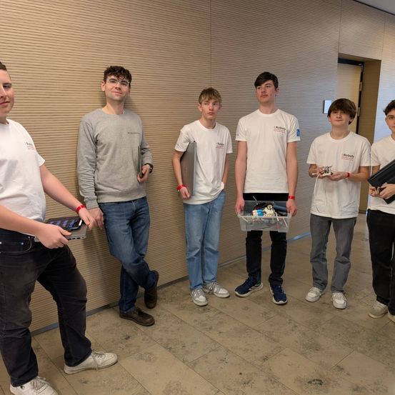 Sechs junge Männer stehen zusammen und lächeln für ein Foto. Sie tragen weiße Hemden und Jeans. Jeder von ihnen hält ein Stück elektronischer Ausrüstung. Der Boden ist gefliest und hinter ihnen befindet sich eine Holzwand.