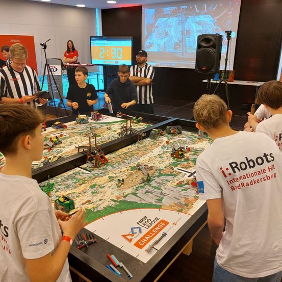 Kinder nehmen an einer First Lego League Challenge mit Robotern auf einem Tisch in einem Raum teil. Eine digitale Uhr und eine Projektionsleinwand sind im Hintergrund.