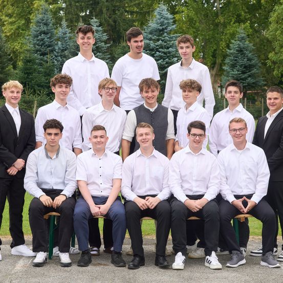 Eine Gruppe junger Männer in weißen Hemden und schwarzen Hosen posiert für ein Foto in einer Freiluftumgebung mit Bäumen im Hintergrund. Einige sitzen auf Bänken, während andere dahinter stehen.