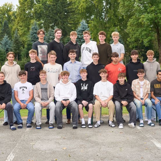 Eine Gruppe junger Männer posiert für ein Foto in einer Außenumgebung, in lässiger Kleidung. Bäume sind im Hintergrund sichtbar.