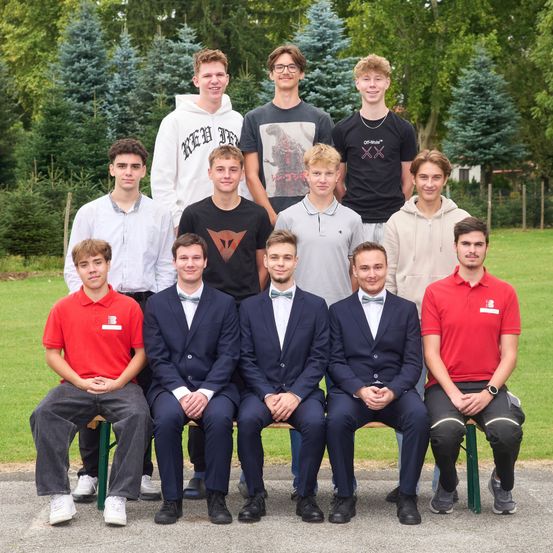 Eine Gruppe junger Männer posiert für ein Foto im Freien. Einige stehen, andere sitzen auf einer Bank. Sie tragen eine Mischung aus lässiger und formeller Kleidung. Hinter ihnen bietet eine Rasenfläche mit Bäumen einen natürlichen Hintergrund.