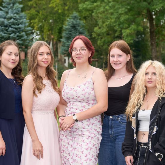 Fünf junge Frauen stehen lächelnd für ein Foto in einem Park. Jede Frau trägt verschiedene Kleidungsstücke, von lässig bis schick. Sie stehen vor üppigen grünen Bäumen, mit Sonnenlicht, das die Szene erhellt.