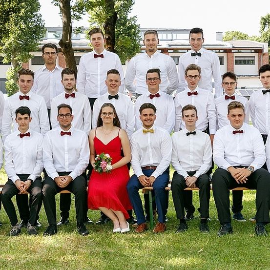 Eine Gruppe von Menschen, die weiße Hemden und Fliegen tragen, sitzen auf Bänken auf einem Rasenfeld, mit einer Frau in einem roten Kleid in der Mitte.
