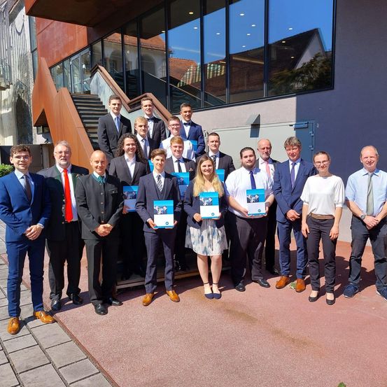 Eine Gruppe von Männern und Frauen in formeller Kleidung steht vor einem Gebäude und hält Urkunden. Die Gruppe besteht aus einer Frau in einem Kleid, zwei Männern in Krawatten und mehreren anderen in Anzügen.