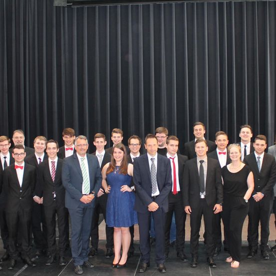 Eine Gruppe formell gekleideter Personen steht vor einem schwarzen Vorhang, möglicherweise für ein Teamfoto. Sie tragen Anzüge und Krawatten, einige tragen Fliegen. Die Frau in der Mitte lächelt.