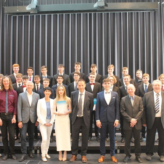 Eine Gruppe formell gekleideter Personen posiert für ein Foto auf einer Bühne mit schwarzem Vorhang als Hintergrund.