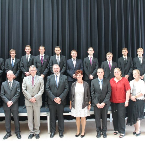 Eine Gruppe von Personen in formeller Kleidung, die vor einem schwarzen Vorhang stehen, möglicherweise für ein Foto. Die Männer tragen Anzüge und Krawatten, während die Frauen Kleider tragen. Sie alle lächeln und scheinen für ein Foto zu posieren.