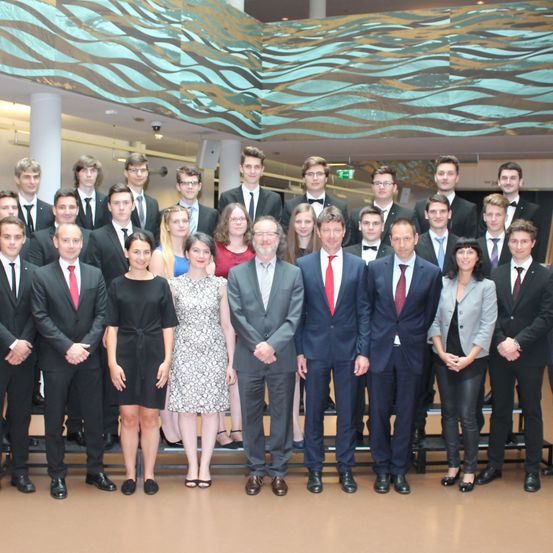 Eine Gruppe formell gekleideter Personen steht gemeinsam für ein Foto, wahrscheinlich bei einer Veranstaltung. Sie tragen Anzüge und Krawatten, einige Frauen sind in Kleidern. Ein Mann in der Mitte hält ein Mikrofon.