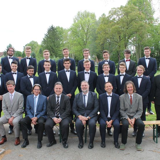 Eine Gruppe formell gekleideter Personen, darunter einige mit Fliege, posiert für ein Foto auf einer Bank in einem Park mit Bäumen im Hintergrund.