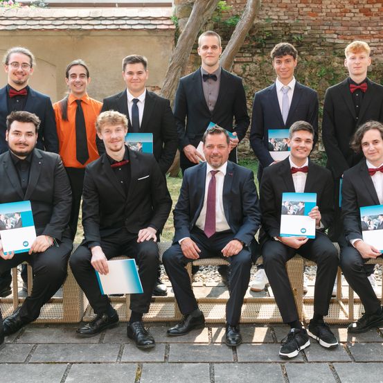 Eine Gruppe formell gekleideter junger Männer in Anzügen und Fliegen sitzen und stehen, halten Bücher und lächeln in die Kamera.