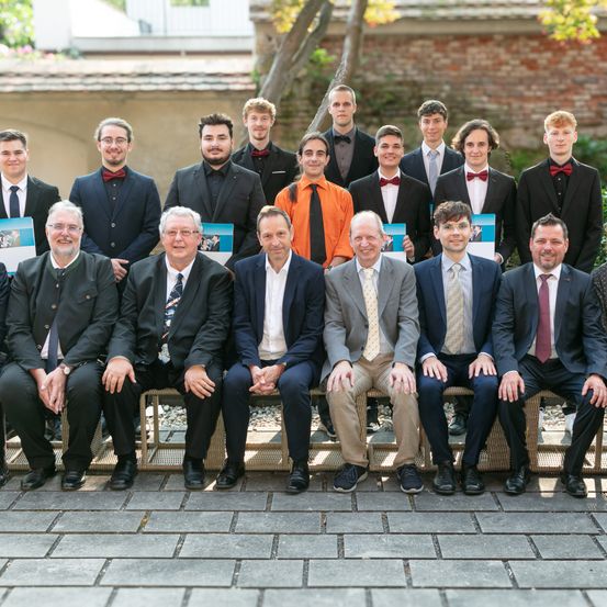 Eine Gruppe von Männern und Jungen, alle in Anzügen und Krawatten gekleidet, posiert für ein Foto im Freien. Einige der Jungen halten Urkunden in den Händen.