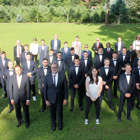 Eine Gruppe formell gekleideter Männer und eine Frau posiert für ein Foto auf einer Rasenfläche mit Bäumen im Hintergrund.