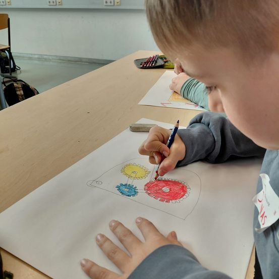 Ein junger Junge sitzt an einem Schreibtisch und malt eine Zeichnung einer Maschine mit Buntstiften aus. Er ist von anderen Kindern und Schulsachen umgeben.