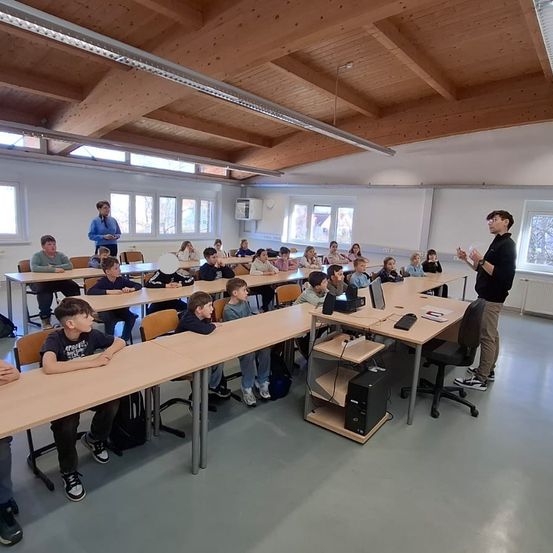 Ein Klassenzimmer mit Holztischen und Stühlen. Schüler sitzen, während ein Mann vorne steht, möglicherweise unterrichtet. Eine Tafel ist an der Wand, und es gibt Fenster an der Seite.