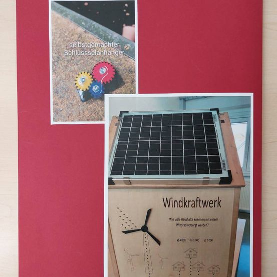 Ein Lehrposter mit einem selbstgemachten Schloss und einer Windturbine mit Solarmodulen wird auf rotem Hintergrund gezeigt. Der Text in Deutsch fragt, wie viele Häuser mit einer Windturbine versorgt werden können.