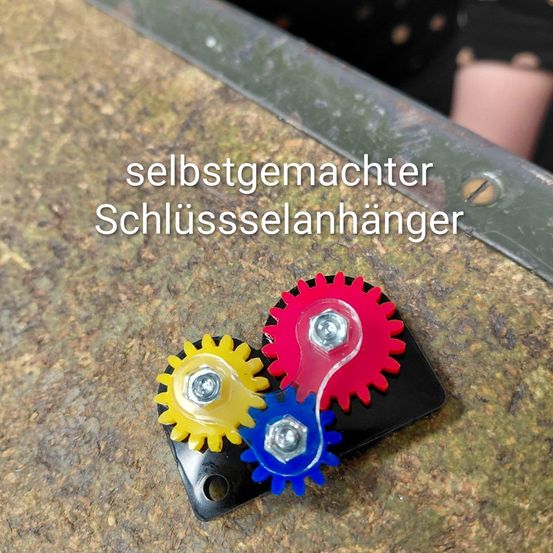 Drei farbige Zahnräder befinden sich auf einer schwarzen Platte mit dem Wort "selbstgemachter Schlussselanhanger" im Bild.