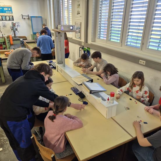 In einer Werkstatt arbeiten Kinder und Erwachsene an einem Projekt. Ein Beamer ist auf einem Ständer montiert. Werkzeuge und Materialien sind auf dem Tisch verteilt.