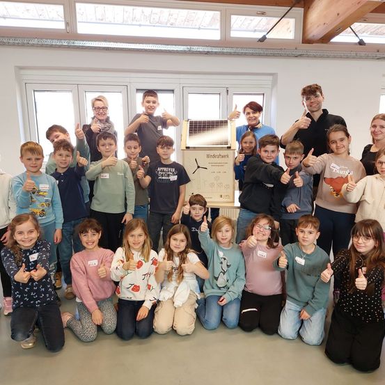 Eine Gruppe von Kindern und Erwachsenen posiert für ein Foto in einem Raum mit einem Solarpanel auf einem Ständer. Sie alle lächeln und zeigen Daumen hoch.